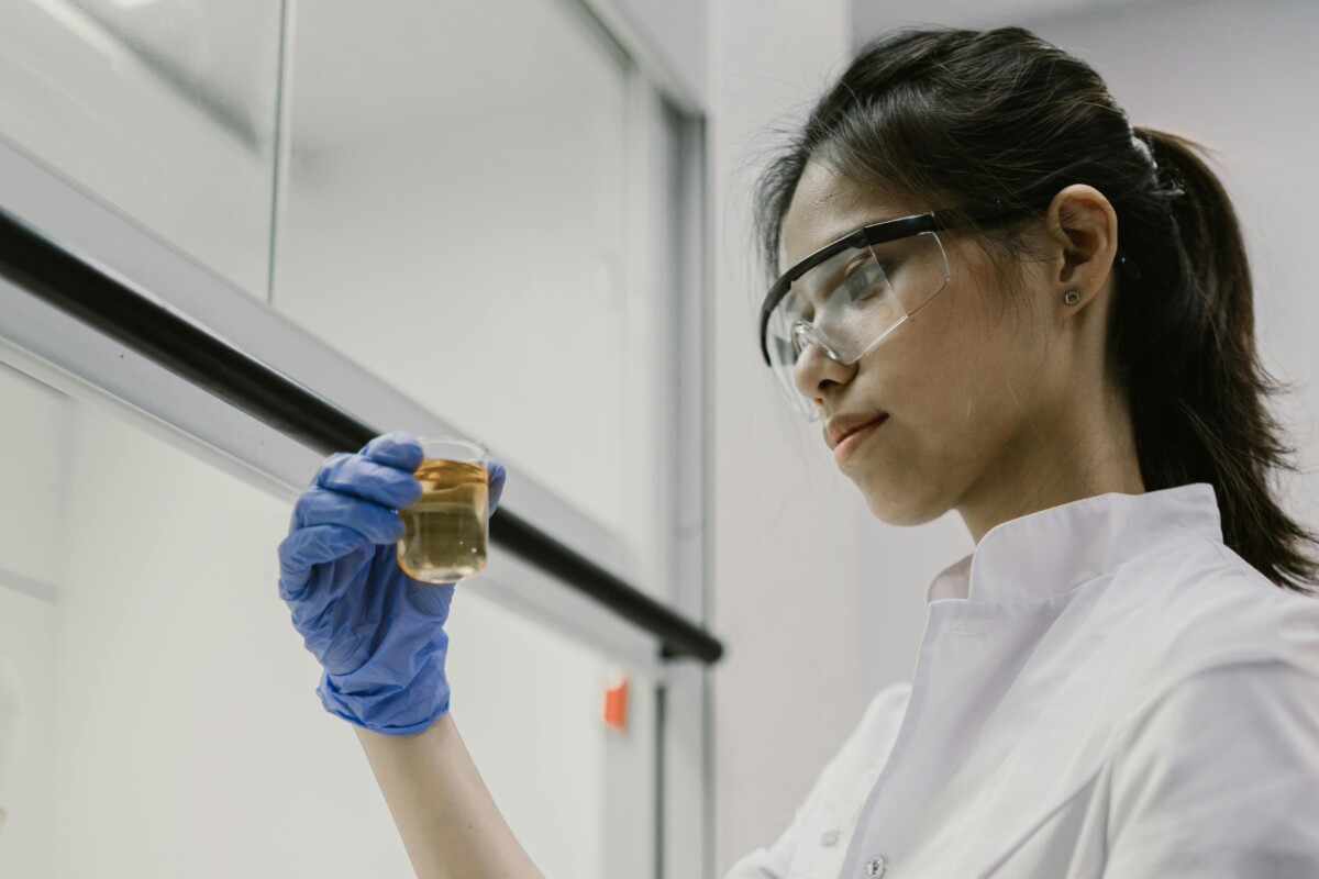 A young woman in lab coat and safety glasses looks at a small beaker of liquid. By Mikhail Nilov on Pexels