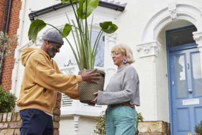 A blonde-haired woman in glasses hands over a big pot plant to a smiling man in a hat and hoodie outside a house that's painted white and has a light blue door.