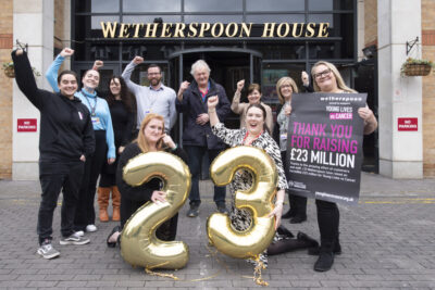 JD Wetherspoon staff cheer outside Wetherspoon House, with gold balloons spelling out 23 - for £23mn raised for Young Lives vs Cancer