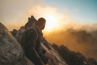 A man sits at the top of a mountain looking at the view, as the sun sets. By Darren Tiumalu on pexels