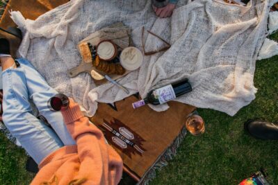 Looking down at a picnic blanket with food and wine on it, and people sitting on it. By Rachel Claire on pexels