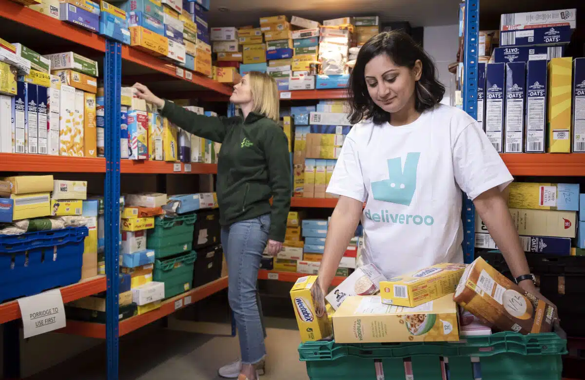 A young woman in a Deliveroo t-shirt holds a tray of food in a Trussell Trust foodbank larder