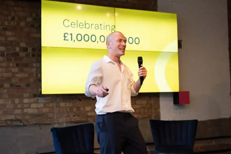 Dan Garrett, CEO of Farewill, speaking at an event to mark £1 billion raised in legacy pledges for charities. He stands in front of a yellow screen saying 'celebrating £1 billion'