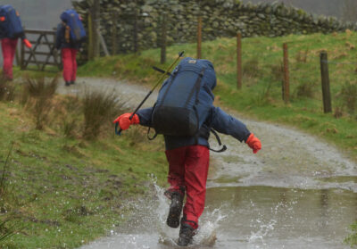 A child in red waterproof trousers with a big backpack splashes through puddles walking up a track