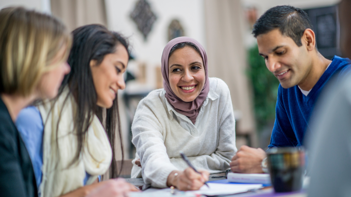 A smiling multicultural group sit at a table. City Bridge Foundation May 24.