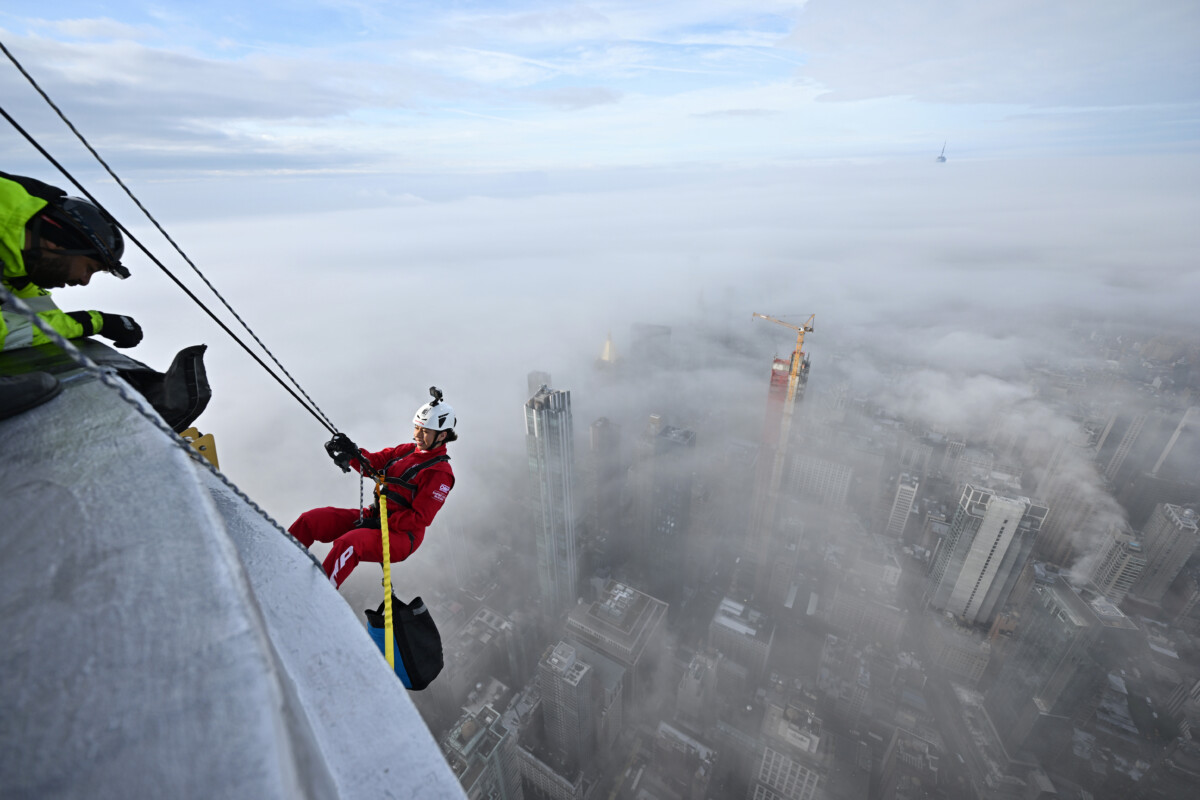 A woman in red suit and white hat starts abseiling down the Empire State Building. Below New York is shrouded in cloud.