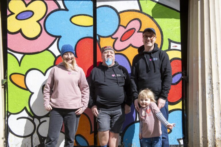 Two men, a woman and a small child pose in front of a colourful mural, wearing clothes from the new Framework Fashion range. Credit Tracey Whitefoot