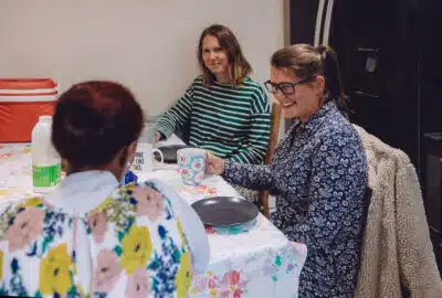 Three women sit around table chatting with plates and mugs. They are from the charity Ella's house