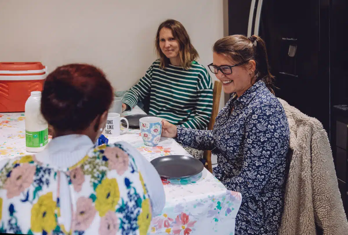 Three women sit around table chatting with plates and mugs. They are from the charity Ella's house