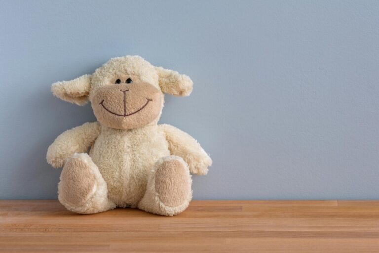 A smiling sheep soft toy sits on a wooden shelf, leaning against a lue grey wall. By Tetyana Kovyrina on pexels