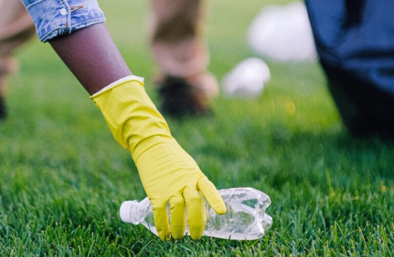 A hand in a yellow glove reaches for an empty plastic bottle lying on some grass. By Anna Shvets on Pexels