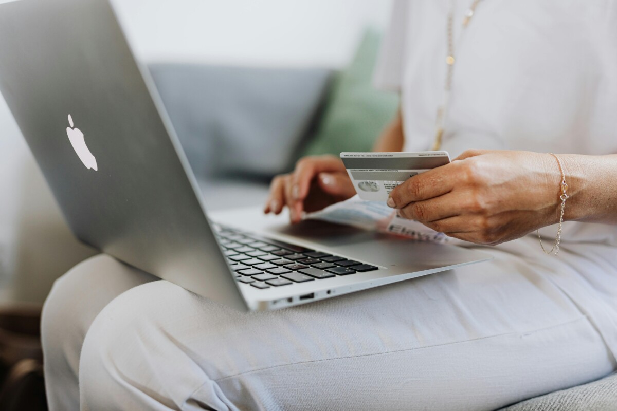 A woman with a laptop on her lap holds her credit card as she shops online. By Karolina Kaboompics on pexels