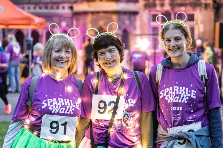 Three women smile at the camera, dressed in Sparkle Walk purple tops with fairy lights and glowsticks.