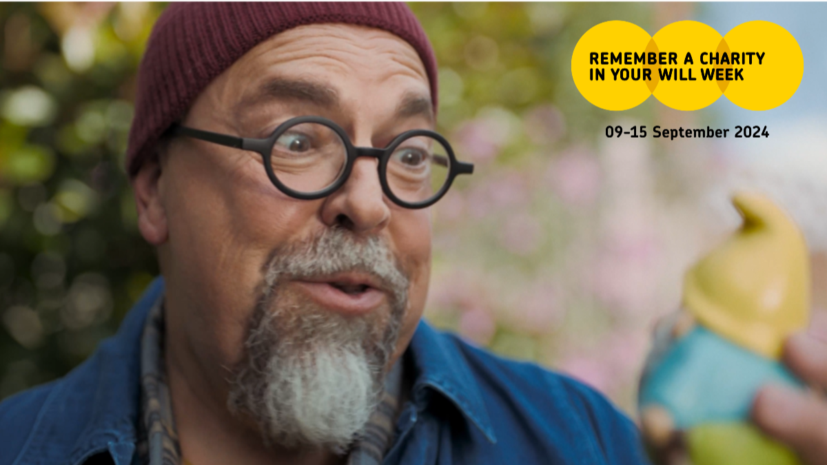 A still from a Remember A Charity Week digital ad showing a man with glasses and a hat looking at a gnome