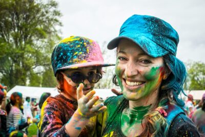 A smiling woman carried a child at a colour run event, both are covered in different colours of paint powder. By Ketan Rajput on Unsplash