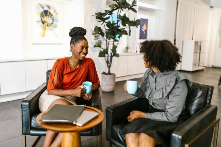 Two women sitting at chairs, talking and smiling over cups of coffee in an office.