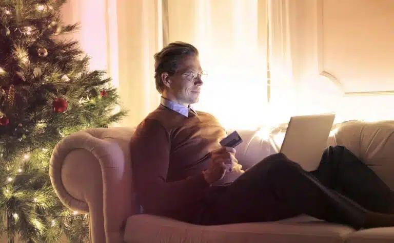 A man sits on his sofa by a Christmas tree, looking at his laptop and holding a credit card. By Andrea Piacquadio on pexels