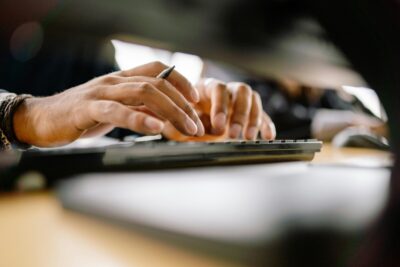 Hands typing on a keyboard, with a pen between two fingers. By Yan Krukov on pexels