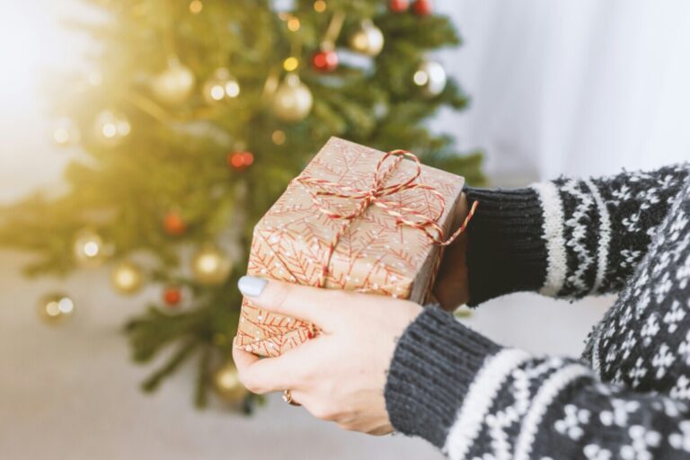 someone in a grey and white patterned sweater handing over a christmas present, in front of a christmas tree. By Jeshoots on unsplash