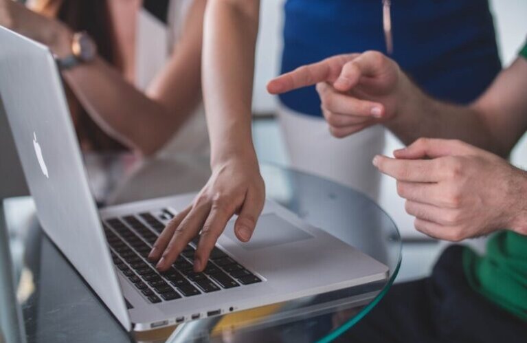 People at a laptop in a meeting, one touches the keys while another points at the screen. By Mimi Thian on Unsplash