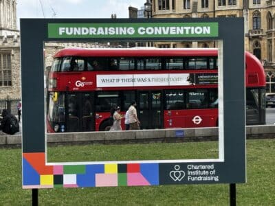 A red London bus is positioned within the frame of a social media frame on the grass outside the venue for the 2025 Fundraising Convention in Westminster, London. The advert on the side of the bus reads "What if there was a fresh start?"