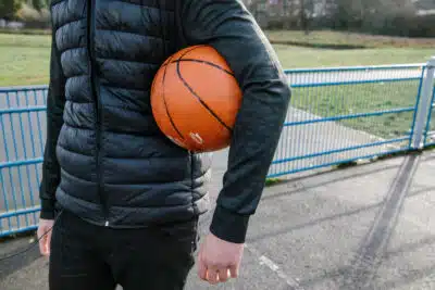 Girl with a basketball under her arm, standing on a concrete basketball court in a park. Close up of ball, torso, hands and puffa-jacket.