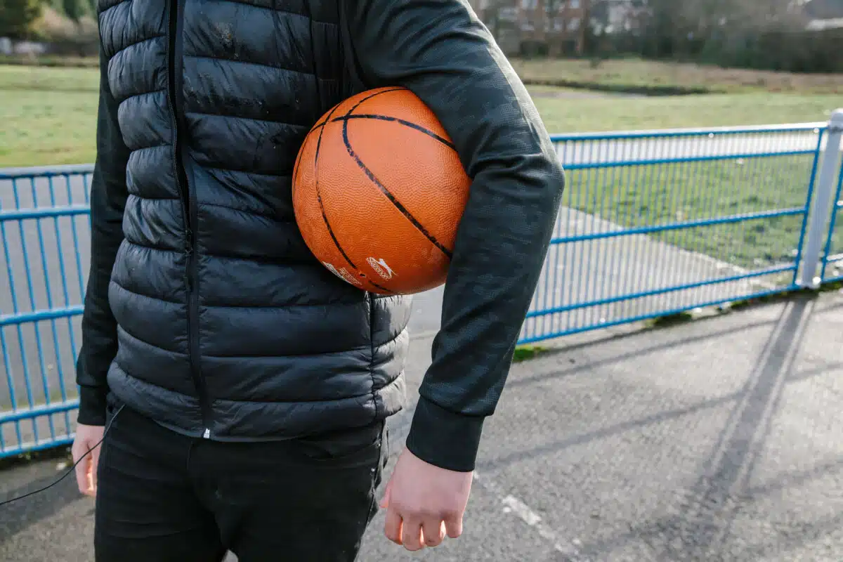 Girl with a basketball under her arm, standing on a concrete basketball court in a park. Close up of ball, torso, hands and puffa-jacket.