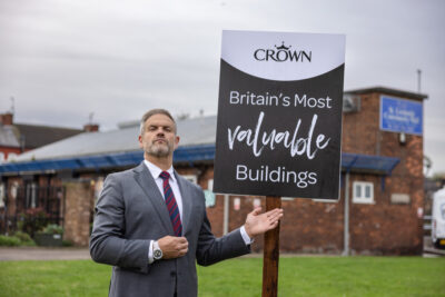 Crown Paints spotlights Britain's most valuable buildings. Man stands beside a mock estate agents' for sale sign.