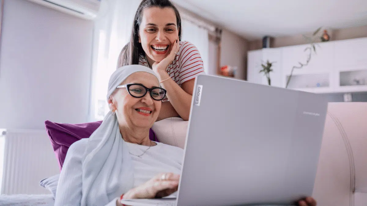 Two women smile as they look at a laptop. The woman sitting down is using the laptop.