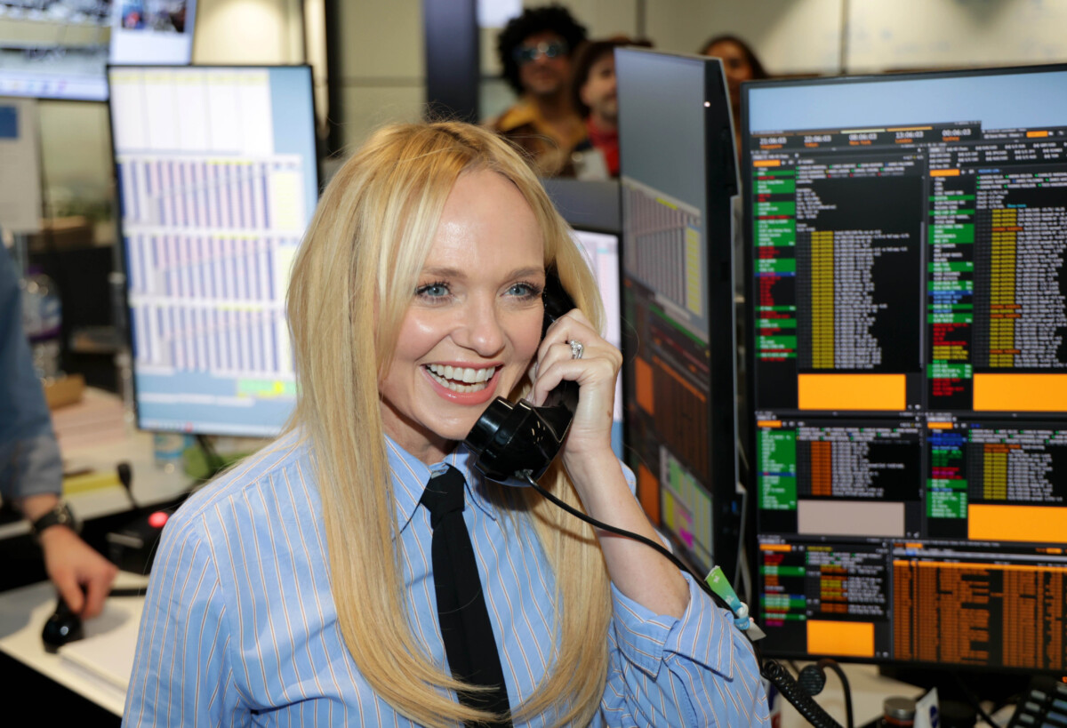Emma Bunton in a blue shirt and black tie smiles while holding a corded phone.